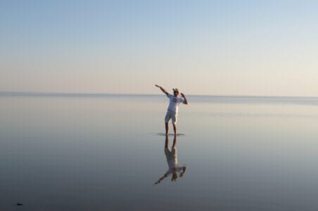 Ein Mensch steht auf einer spiegelglatten Wasseroberfläche, die den wolkenlosen Himmel und die Person perfekt reflektiert. Die Szene vermittelt Ruhe und Weite – ideal für Natur- und Afrikareisen. Die Person trägt ein weißes T-Shirt und Shorts und scheint sich in einer freudigen Pose zu befinden, die an Tanz oder Bewegung erinnert. Dieses Bild symbolisiert die Freiheit und die besondere Atmosphäre, die Reisende auf einer Safari durch Botswana erleben können.